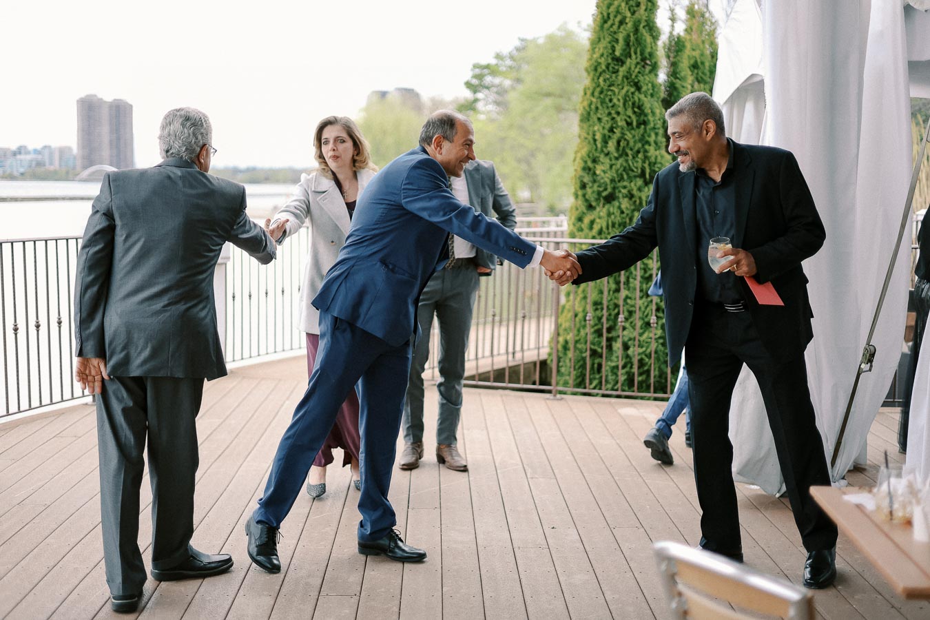 Business professionals shaking hands at an outdoor networking event, with a waterfront and city skyline in the background.