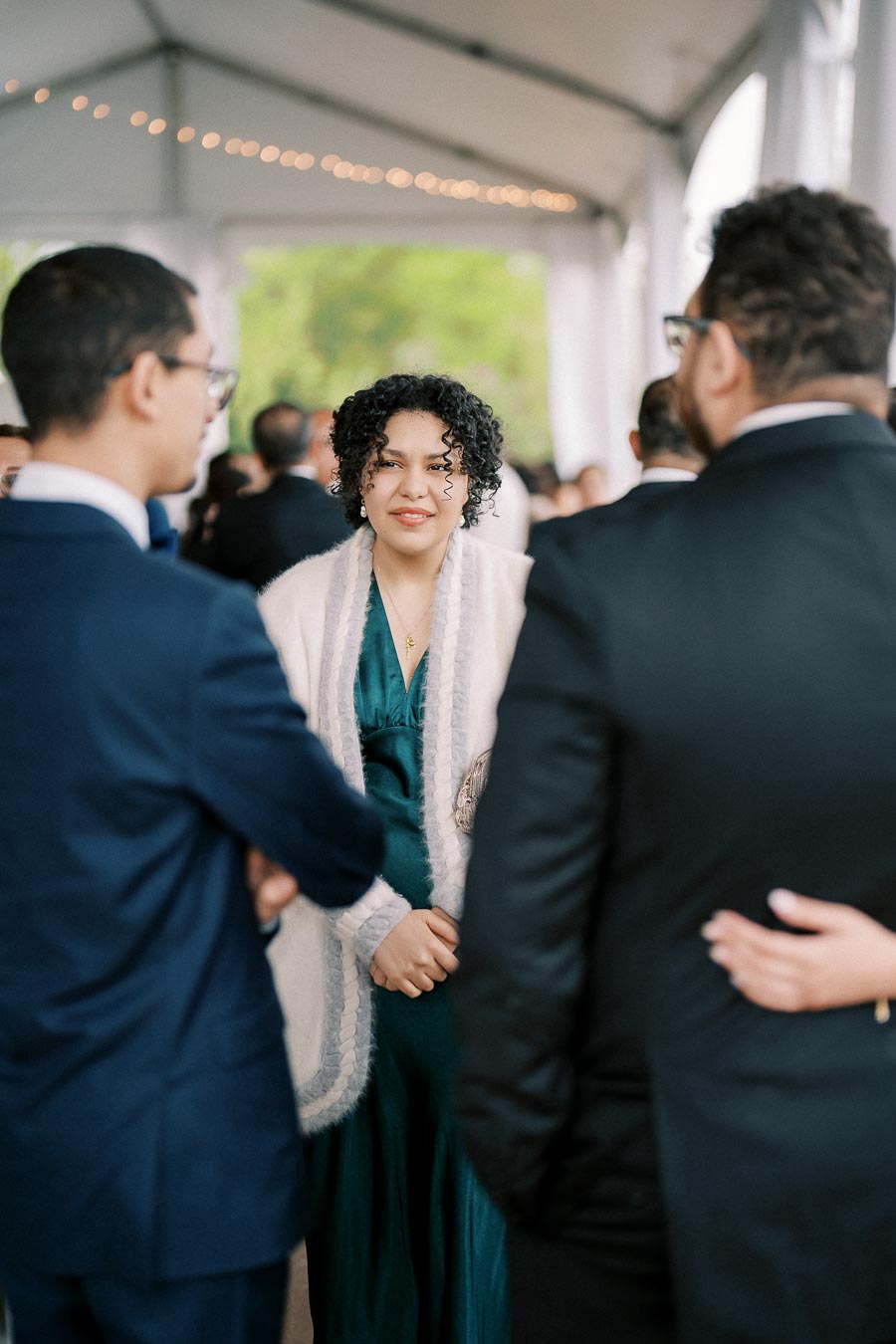 A group of formally dressed individuals interacting at an elegant indoor event, with a focus on a woman in a green dress and light shawl, under a tent with string lights.