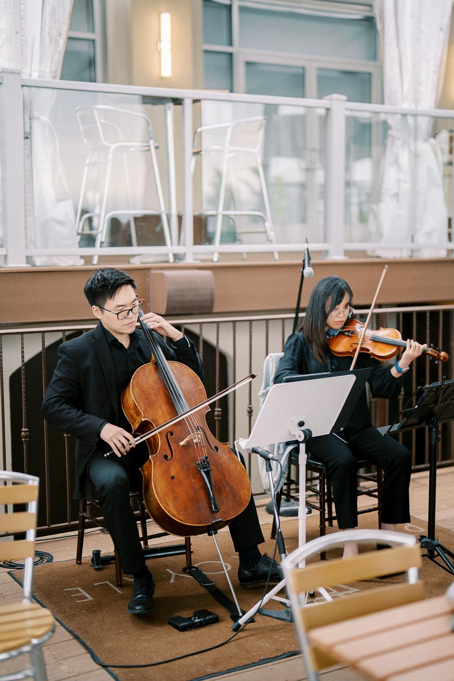 A cellist and violinist in black formal attire perform classical music at an elegant indoor event, with music stands and chairs in the background.