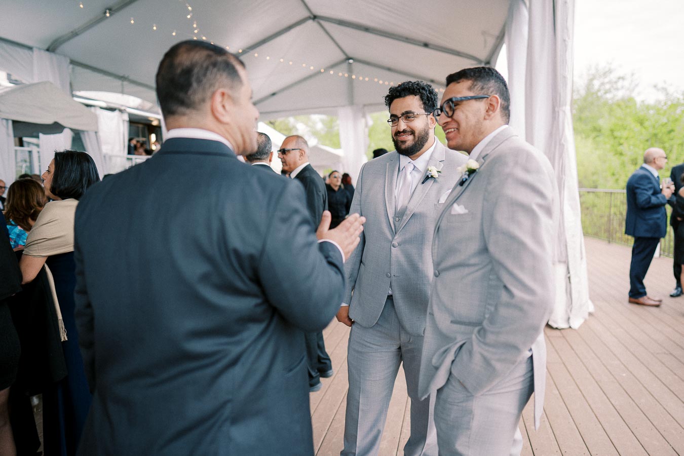 A group of well-dressed men in suits engage in conversation under a tent at a formal outdoor event, with string lights overhead and other guests in the background.