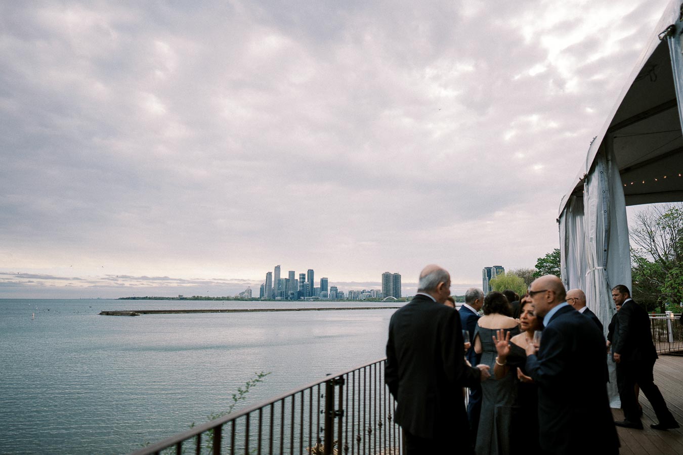 Elevated view of a group of people socializing at a waterfront event, overlooking a lake with a city skyline in the background under a cloudy sky.