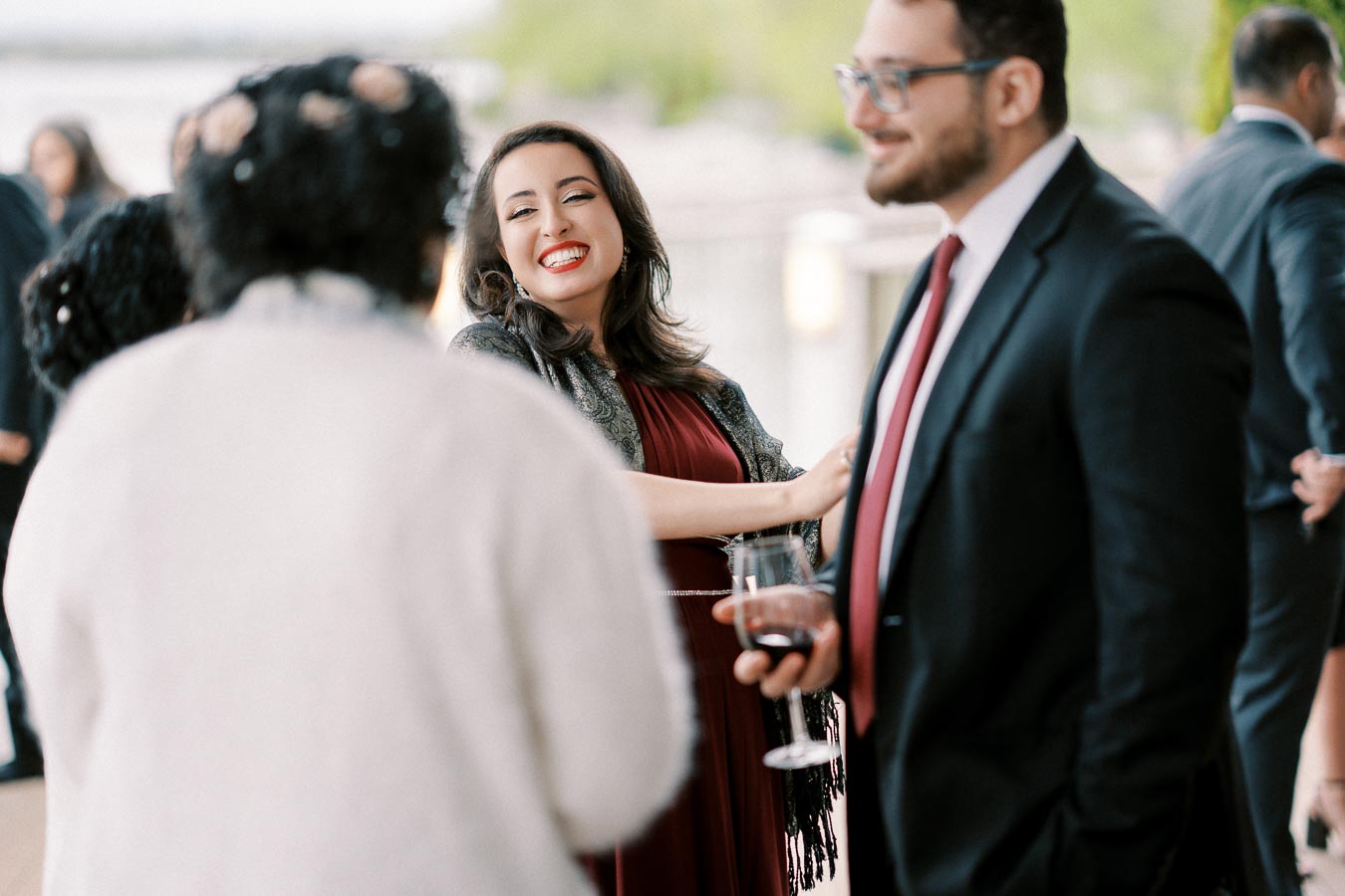 A woman in a red dress smiles while socializing at an outdoor event, surrounded by people in formal attire, holding a glass of wine.