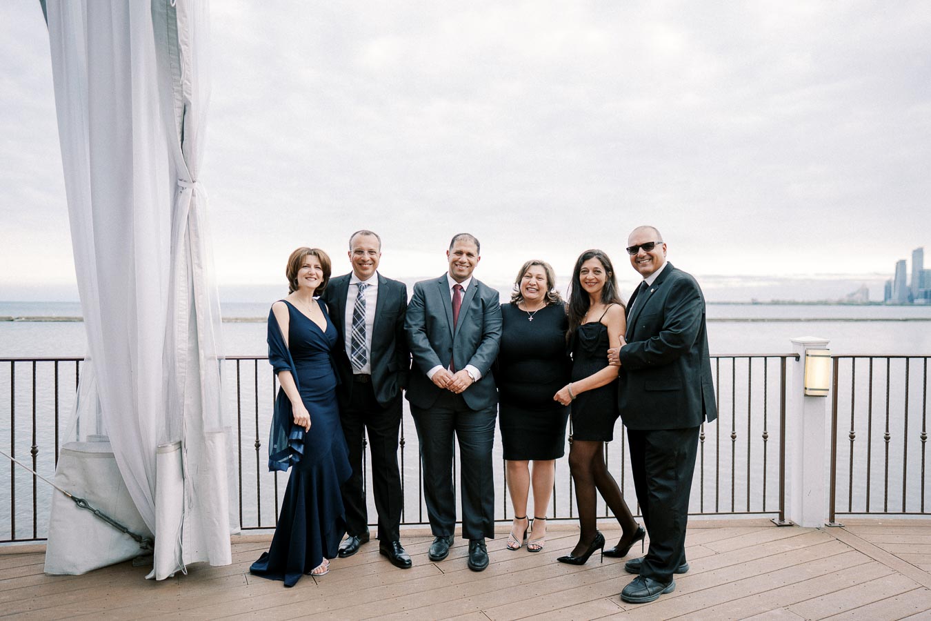A group of six people dressed in formal attire standing on a waterfront deck with a city skyline in the background, under a cloudy sky.
