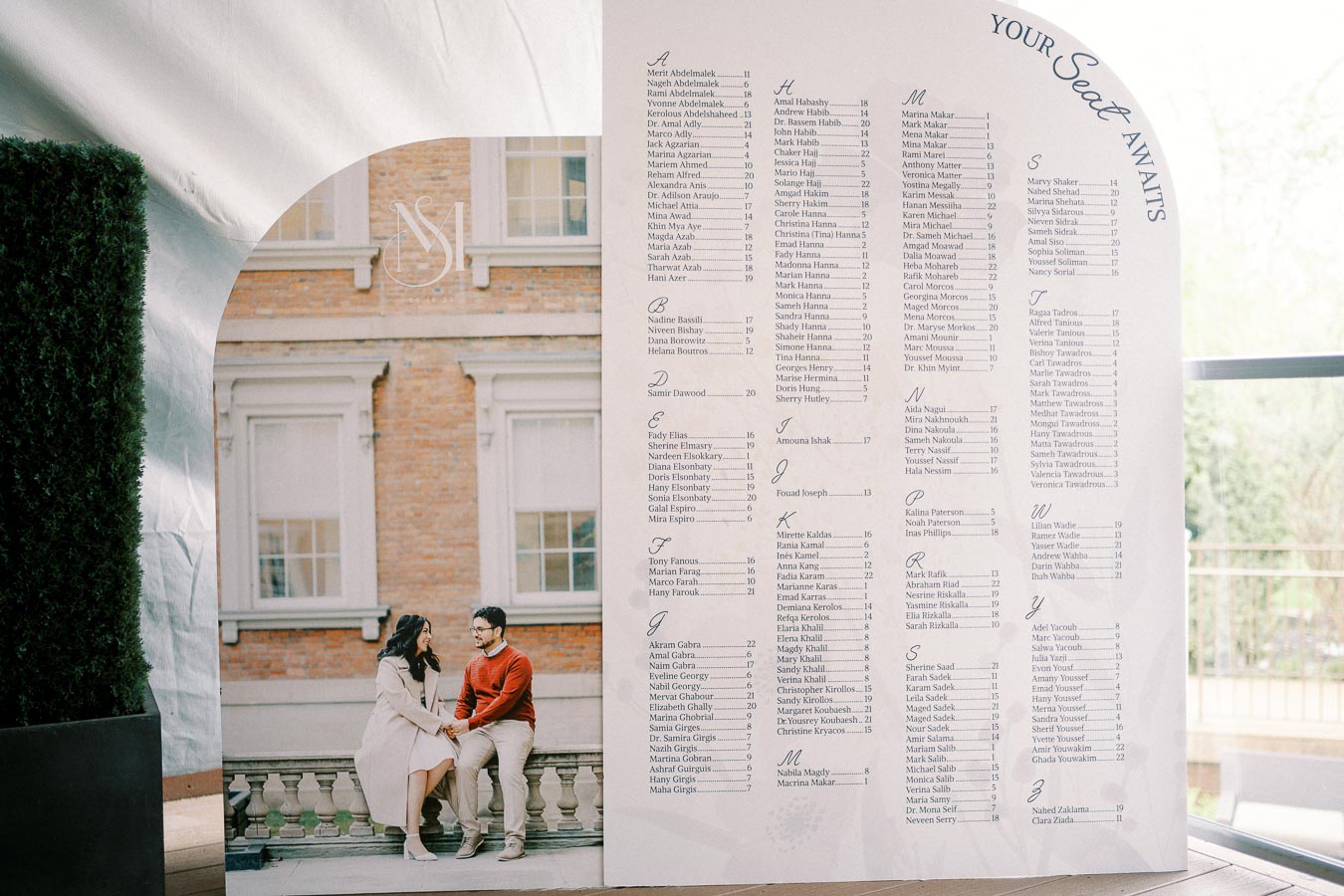 Wedding seating chart display showing guest names alphabetically, with a photo of a couple seated outside a brick building. The chart is labeled Your Seat Awaits, suggesting an elegant and organized wedding event.