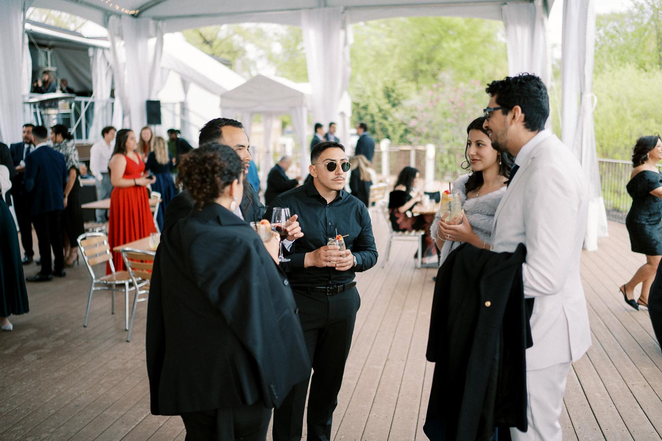 A group of well-dressed people socializing at an outdoor event, some holding drinks and wearing formal attire, with a lively atmosphere and a backdrop of greenery and white event tents.