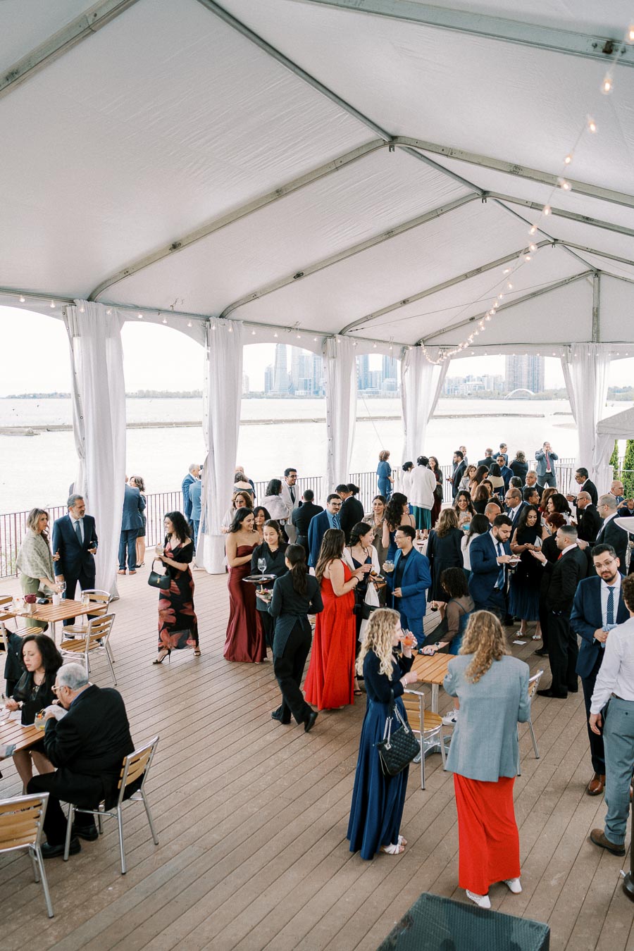 Alt text: Elegant outdoor event under a canopy with a large group of people mingling, wearing formal attire. Waterfront city skyline visible in the background, creating a stylish and scenic atmosphere.