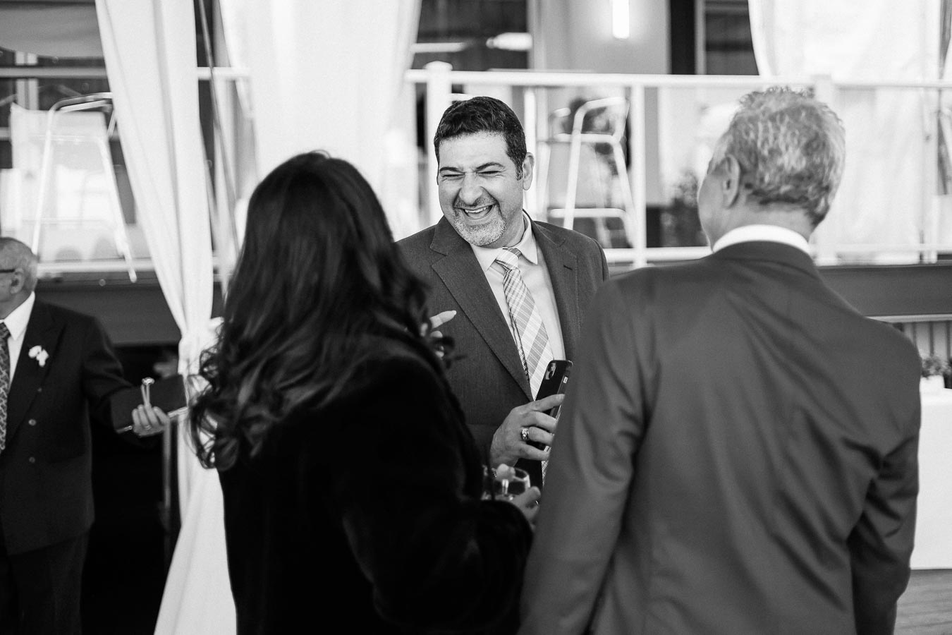 A black and white photo of a group of people in formal attire engaging in a lively conversation at an indoor event. The scene captures their smiling faces and positive energy, with a focus on social interaction and networking.