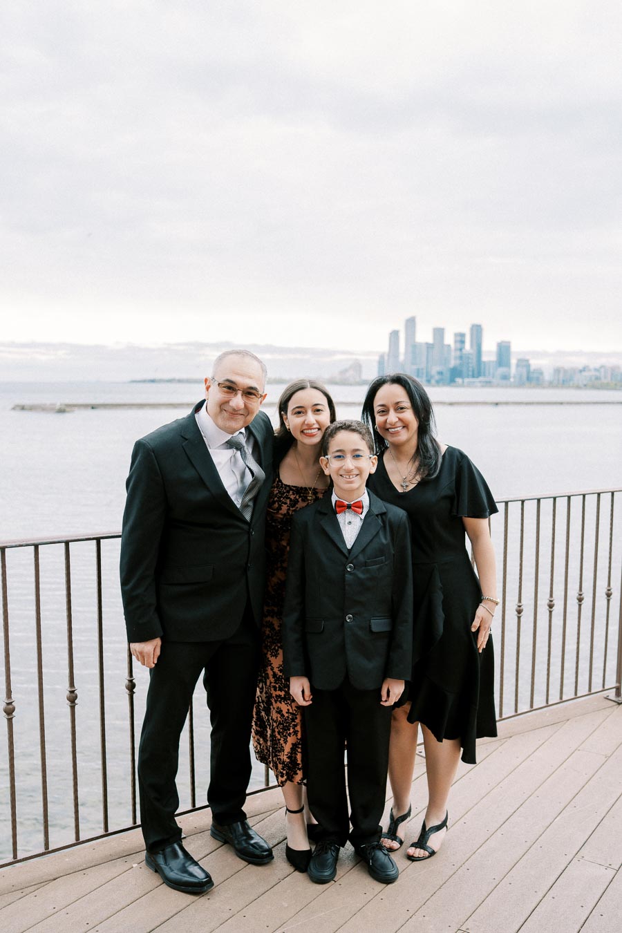 A family of four dressed in formal attire poses on a wooden deck overlooking a serene waterfront, with a cityscape visible in the background.