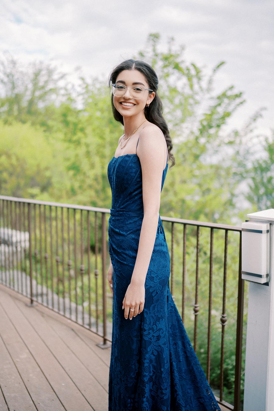 A young woman in a blue lace evening gown standing on a balcony with greenery in the background, smiling and wearing glasses.