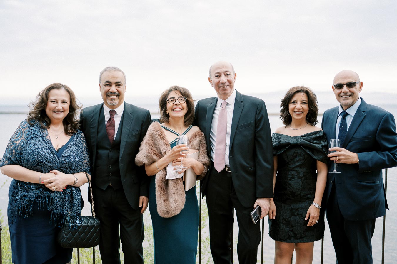 A group of five elegantly dressed adults smiling at a lakeside event, with a scenic waterfront and cloudy sky in the background.