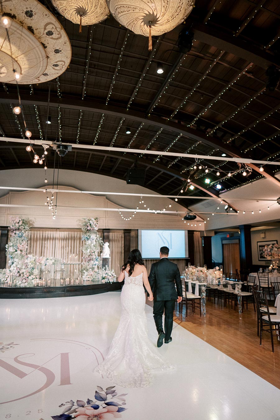 Bride and groom walking hand in hand towards a beautifully decorated wedding reception stage with floral arrangements and elegant lighting in a spacious venue.