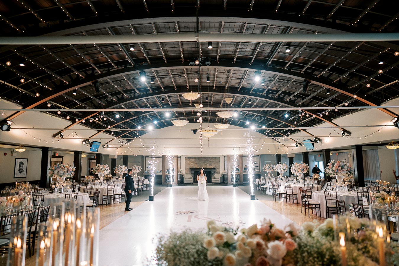 Wedding reception hall with elegant decor, featuring a high ceiling adorned with string lights. A bride and groom stand on a white dance floor surrounded by floral arrangements and lit sparklers, creating a romantic and celebratory atmosphere.