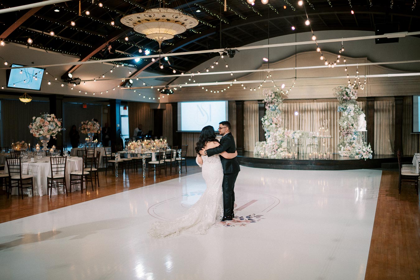 A couple shares their first dance in an elegantly decorated wedding venue, featuring string lights, floral arrangements, and a grand chandelier.