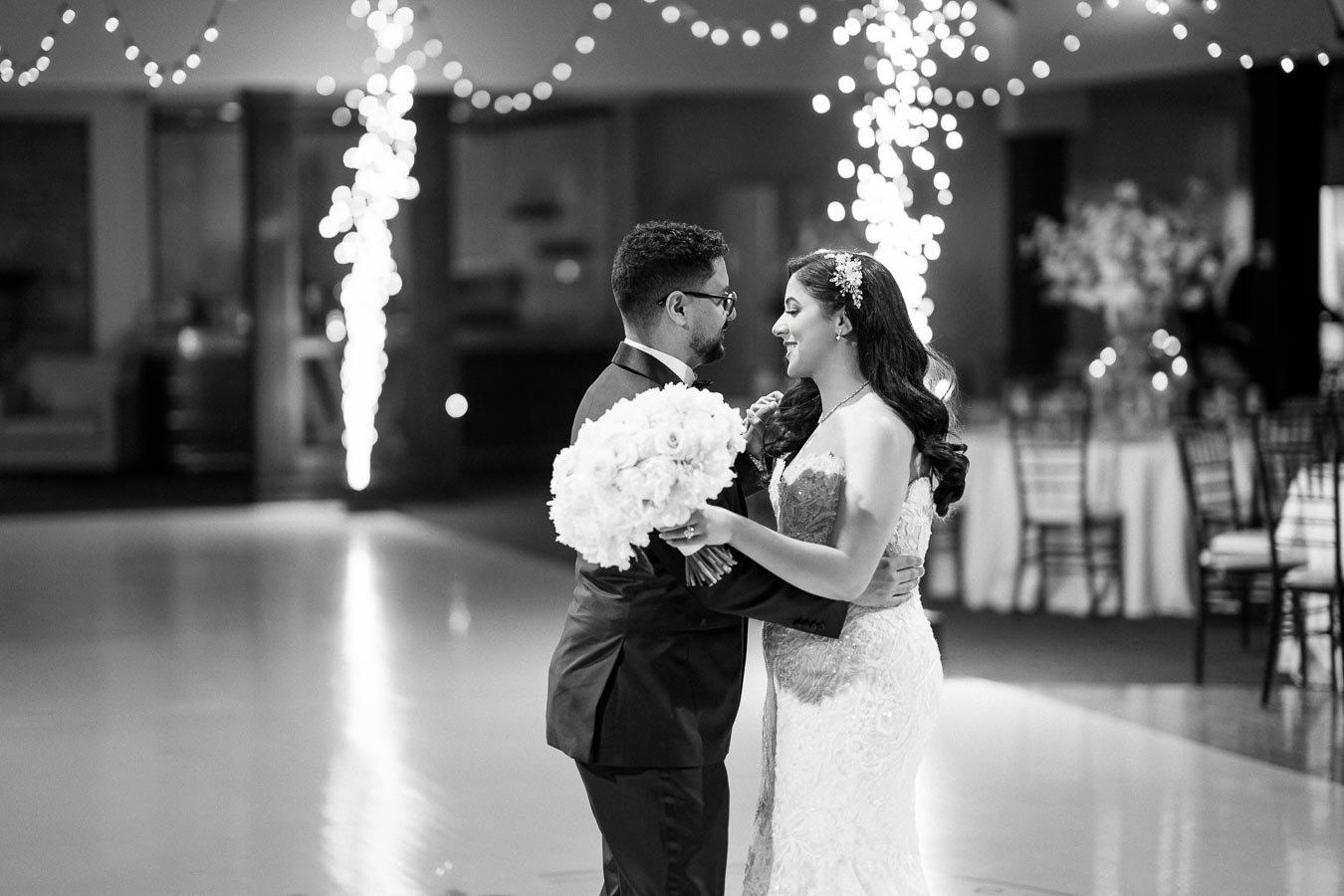 Black and white image of a newlywed couple sharing their first dance at a wedding reception, with the bride holding a bouquet of white flowers and twinkling fairy lights in the background.