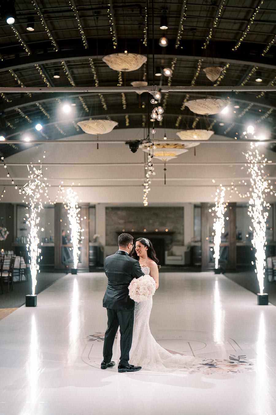 A bride and groom share a romantic first dance on a decorated dance floor lit by indoor sparklers, under a ceiling adorned with hanging lights, creating an enchanting wedding reception atmosphere.