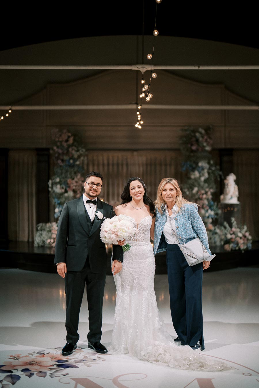 A bride and groom pose with a formally dressed guest at an elegant indoor wedding venue, with floral arrangements and soft lighting enhancing the celebratory atmosphere.