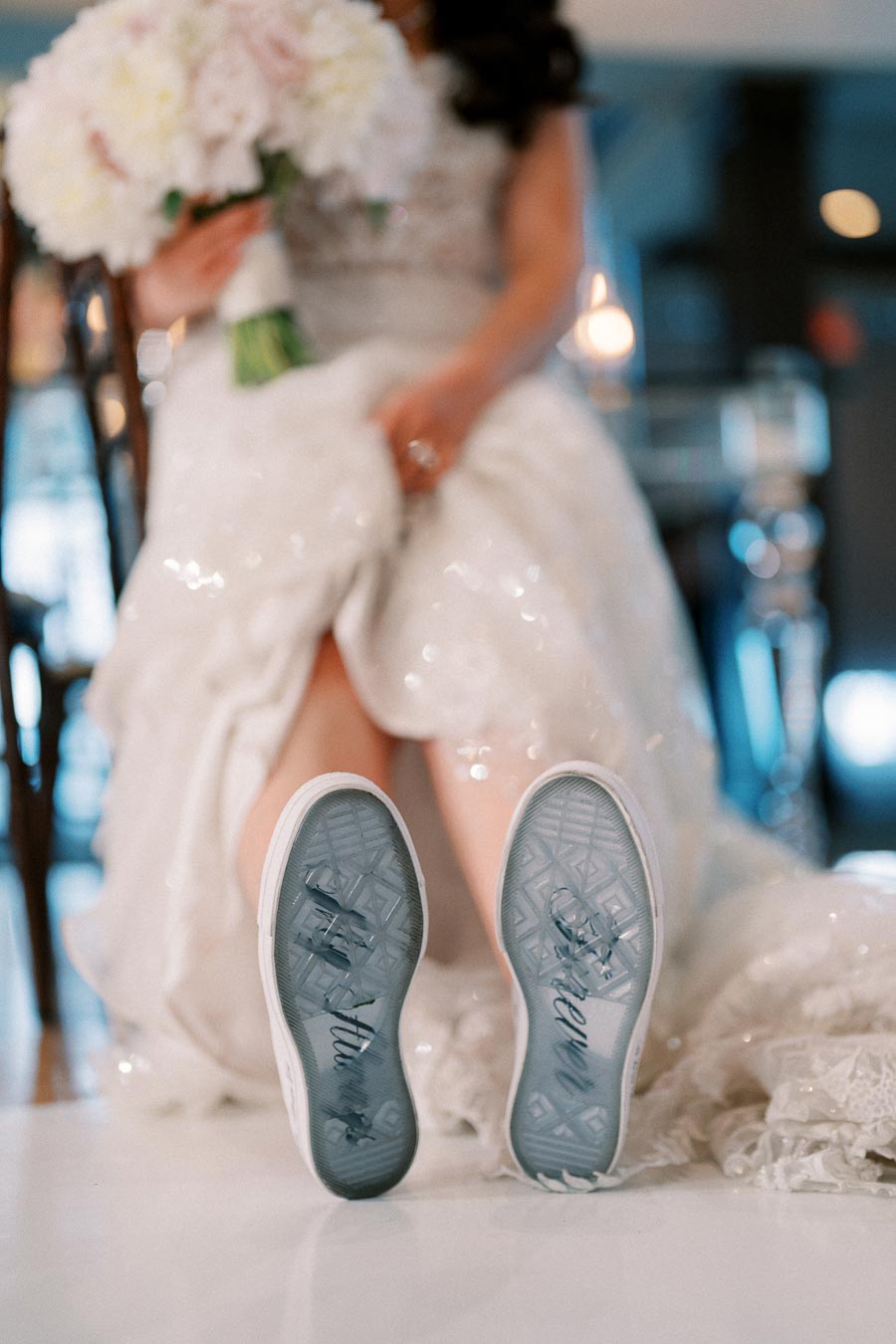 Bridal sneakers with cursive writing on soles, bride holding bouquet in blurred background, showcasing unique wedding style.