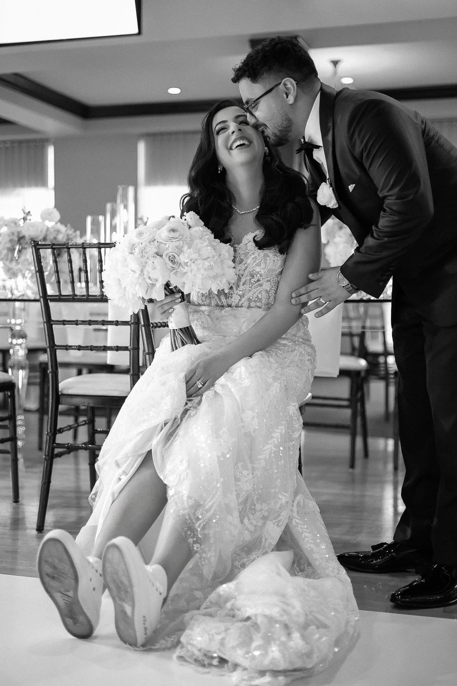 Smiling bride in lace wedding dress sits with bouquet, husband leaning in for a kiss, wearing suit and tie in elegant wedding venue.
