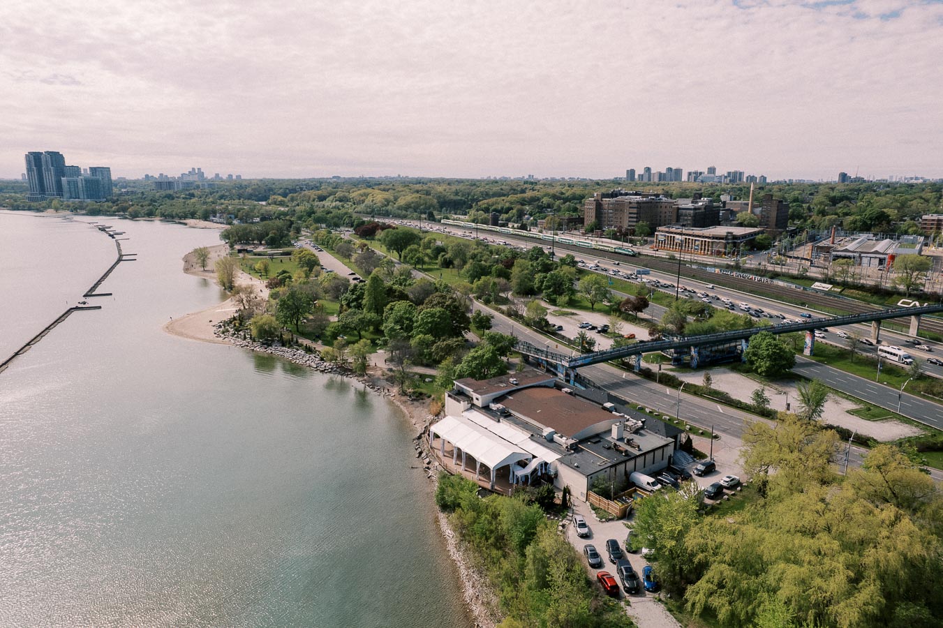 Aerial view of a lakeside urban park with lush greenery, adjacent highways, and a waterfront building, under a cloudy sky.