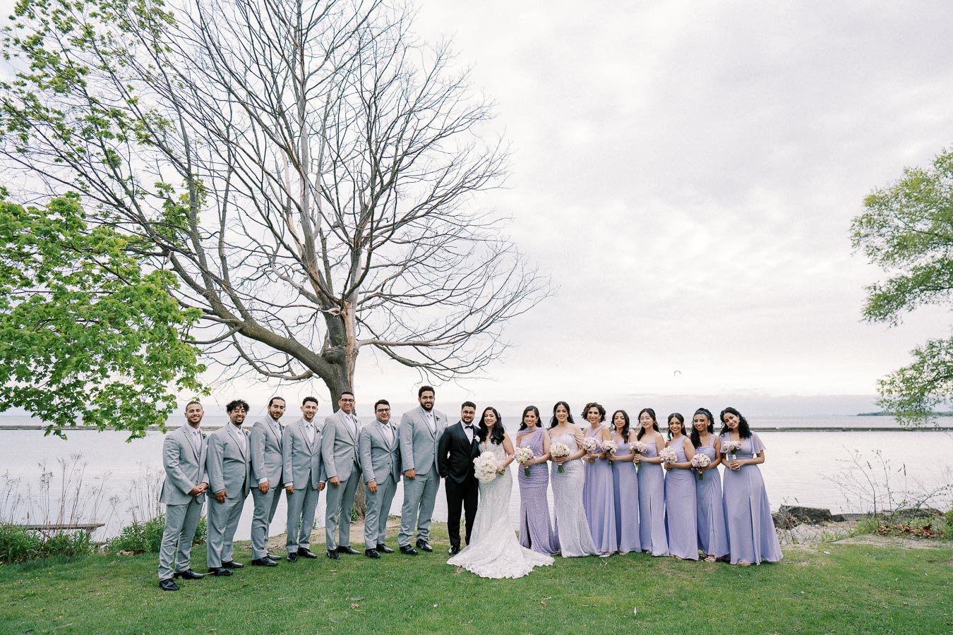 Wedding party by a lakeside, featuring bridesmaids in lavender dresses and groomsmen in gray suits, standing with the couple under a large tree against a serene water backdrop.