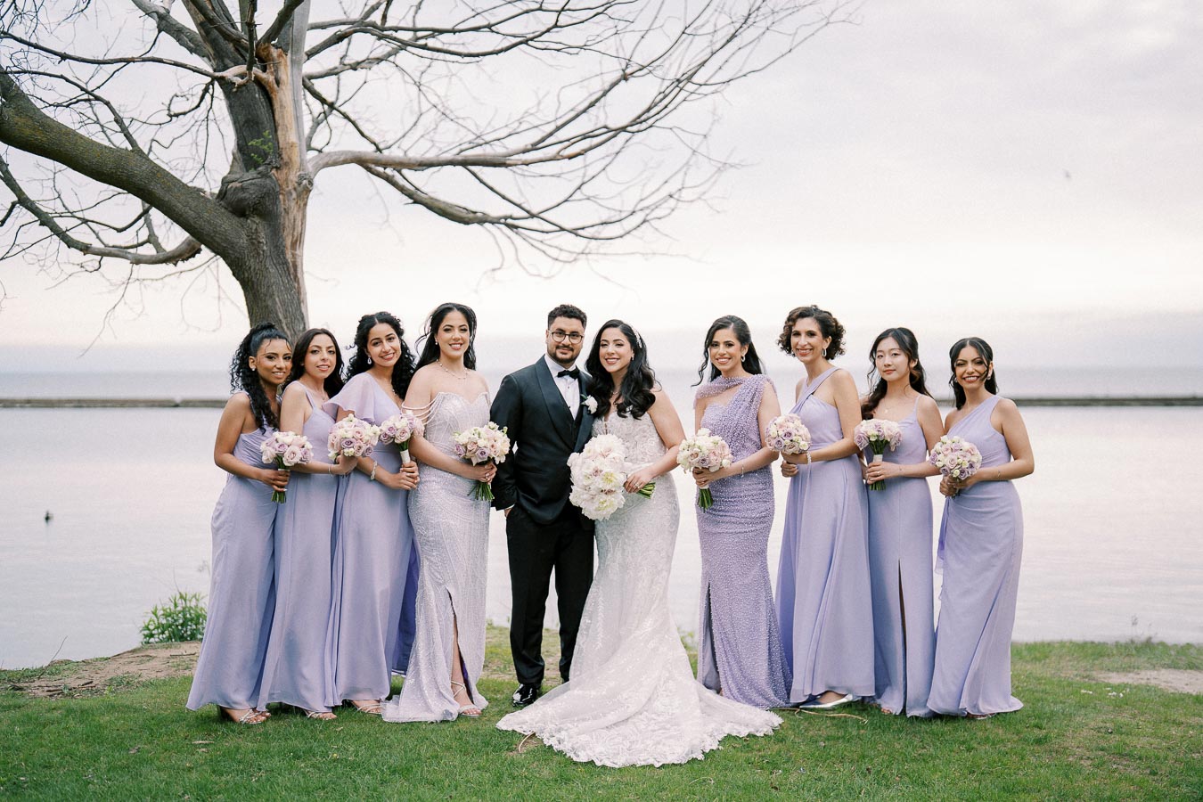 A wedding party posing near a lake with a tree in the background. The bride in a white gown stands beside the groom in a black tuxedo, surrounded by bridesmaids in lavender dresses holding bouquets, under an overcast sky.