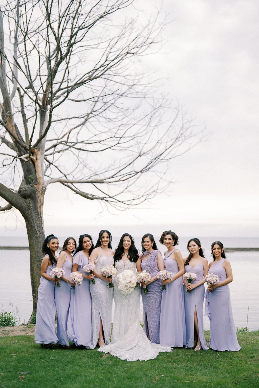 A bride in a white gown stands smiling with her bridesmaids dressed in lavender dresses, each holding bouquets, in an outdoor setting by a lake with a bare tree in the background.