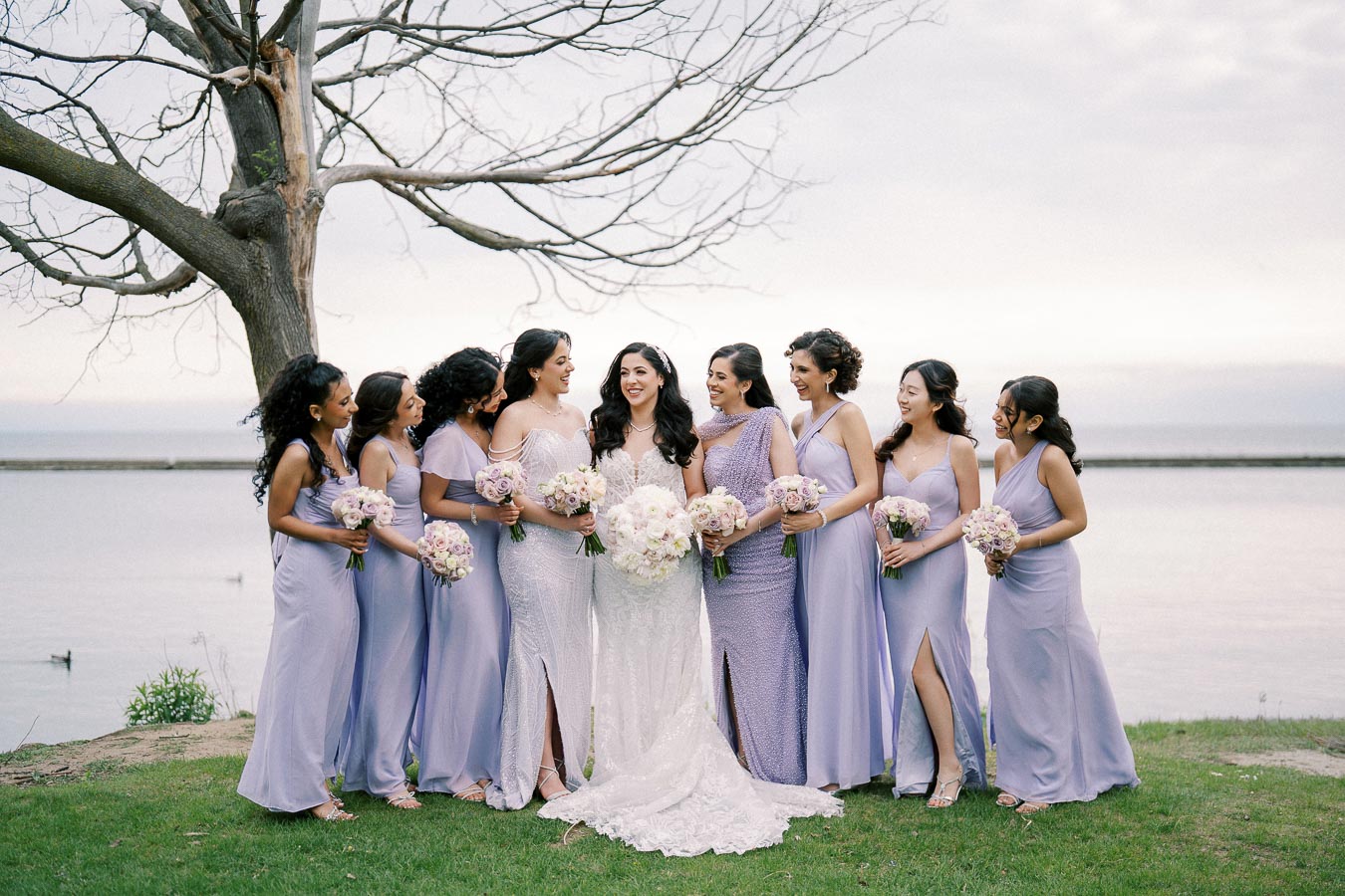 A bride and her bridesmaids in lavender dresses stand together holding bouquets by a lakeside under a bare tree, showcasing a joyful wedding moment.