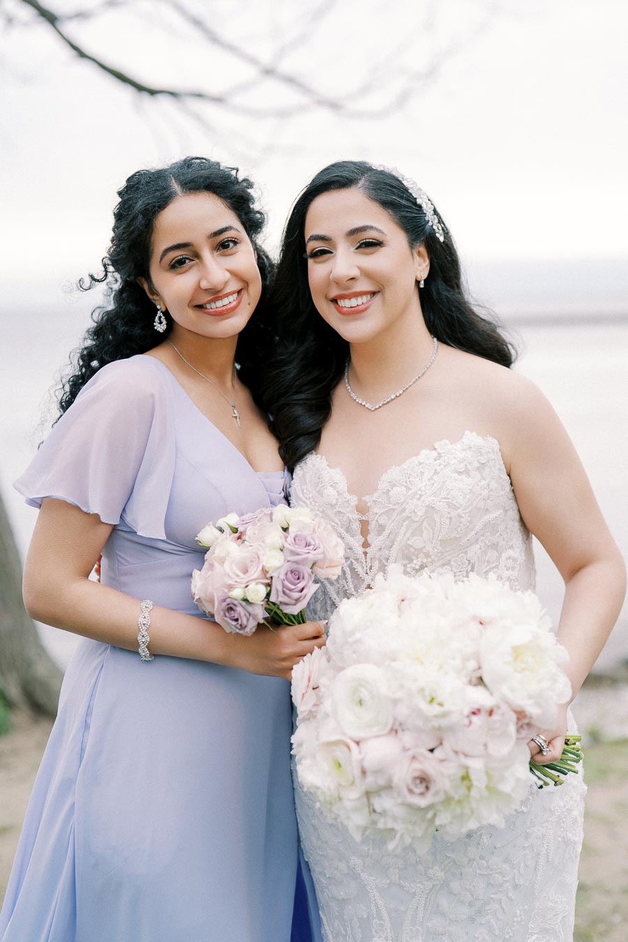Two smiling women at a wedding, with one in a white lace bridal gown holding a bouquet of white and blush flowers, and the other in a lavender bridesmaid dress holding a bouquet of purple roses, standing outdoors.