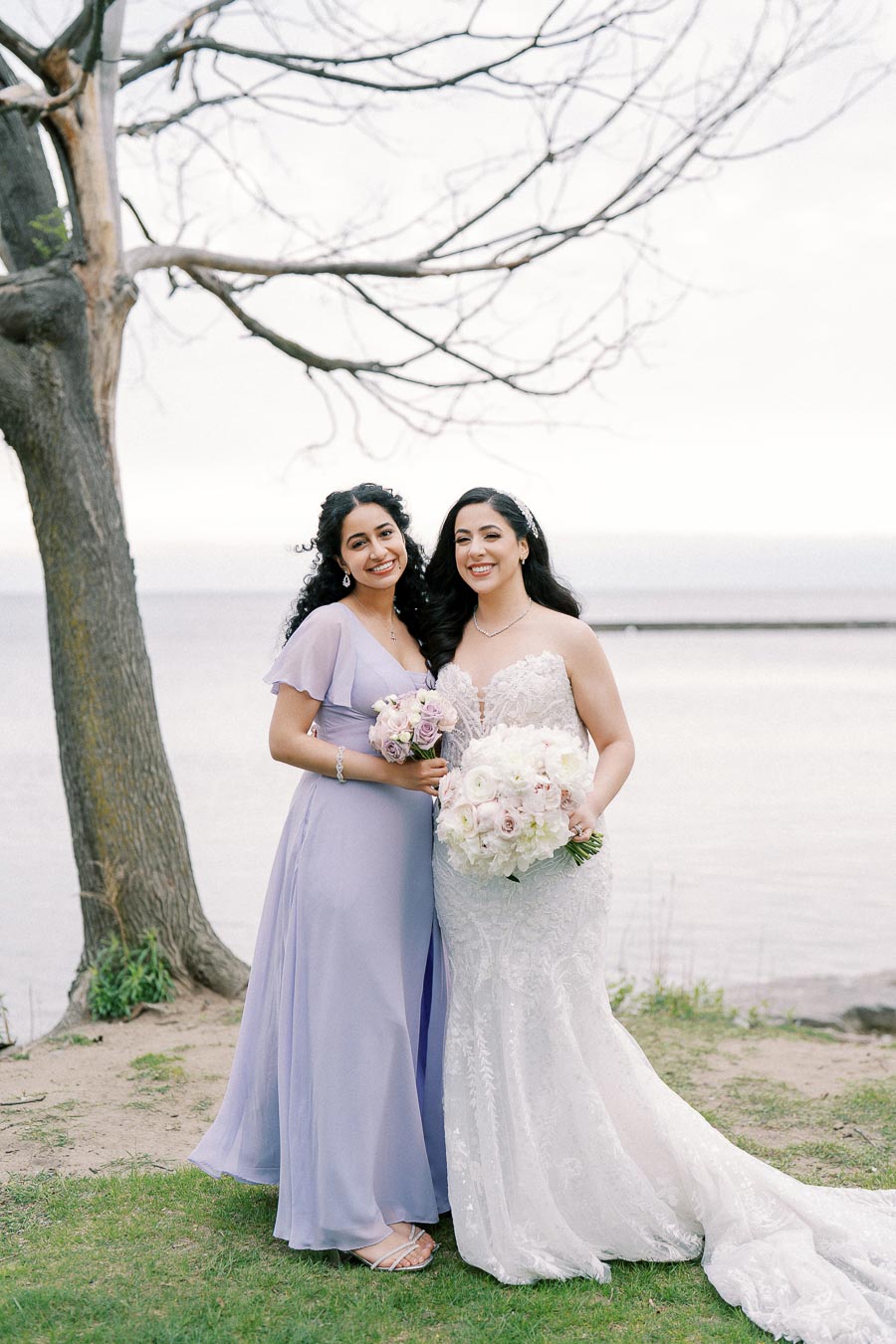 Two women standing together outdoors, one in a wedding gown and the other in a lavender bridesmaid dress, both holding bouquets of flowers with a waterfront and tree in the background.