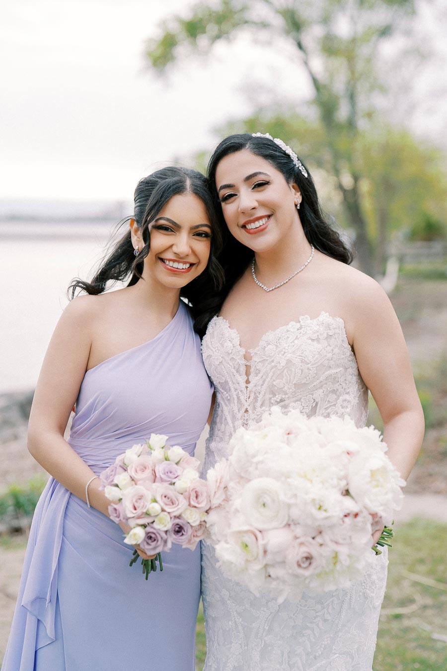 Bride and bridesmaid smiling and holding flower bouquets, with bride in an elegant lace wedding dress and bridesmaid in a lavender gown, outdoor wedding setting.