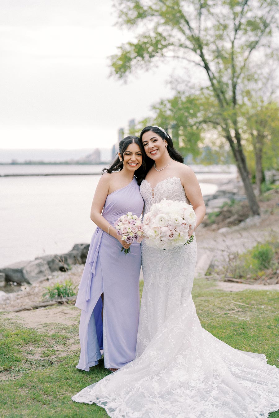 Two women smiling, dressed in formal attire near a lakeside. One is wearing a white lace wedding dress holding a bouquet of flowers, and the other is in a lavender bridesmaid dress with a bouquet. The background features trees and a serene body of water.
