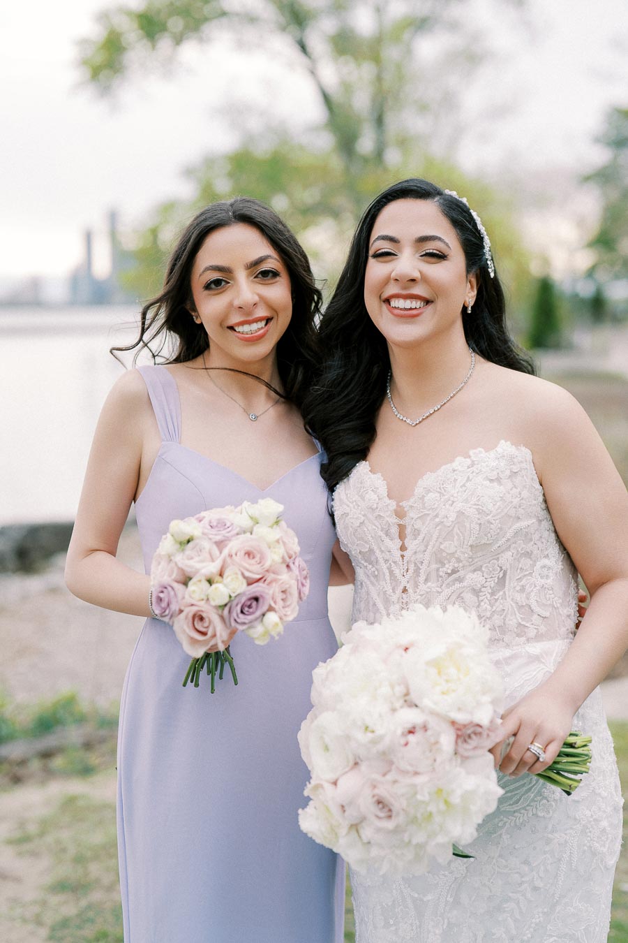 Two women smiling outdoors, one in a white lace wedding dress holding a bouquet of white and pink flowers, and the other in a lavender bridesmaid dress holding a similar bouquet, with greenery and water in the background.
