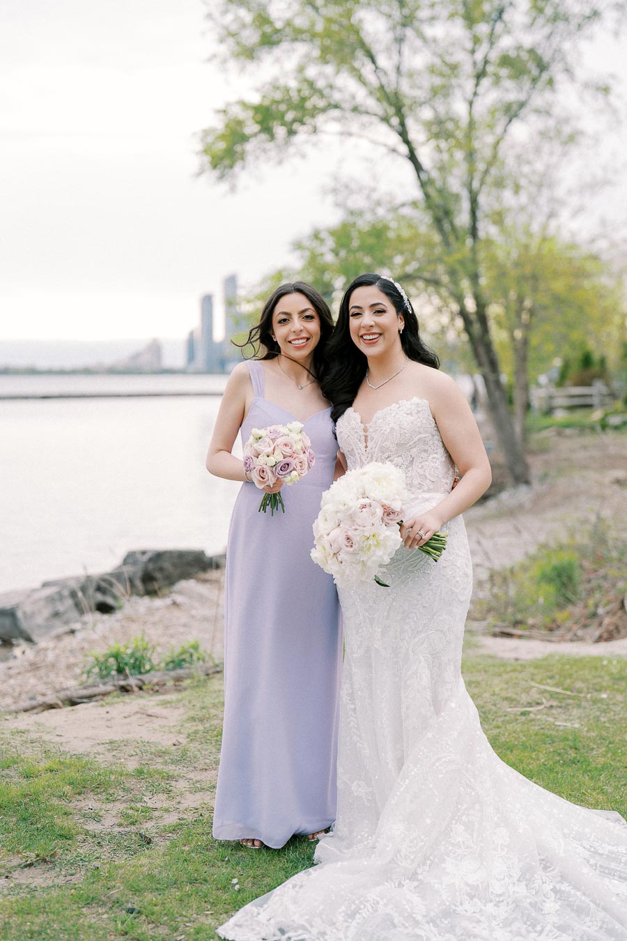 Two women smiling in elegant dresses, holding bouquets, standing outdoors by a waterfront with trees in the background on a wedding day.