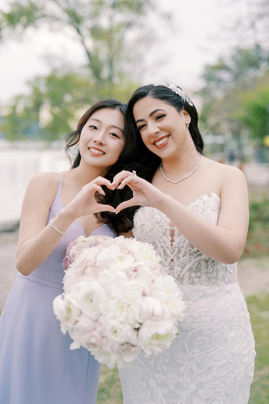 A bride and bridesmaid smiling and forming a heart shape with their hands, holding a bouquet of pink and white flowers, standing outdoors in a park setting.