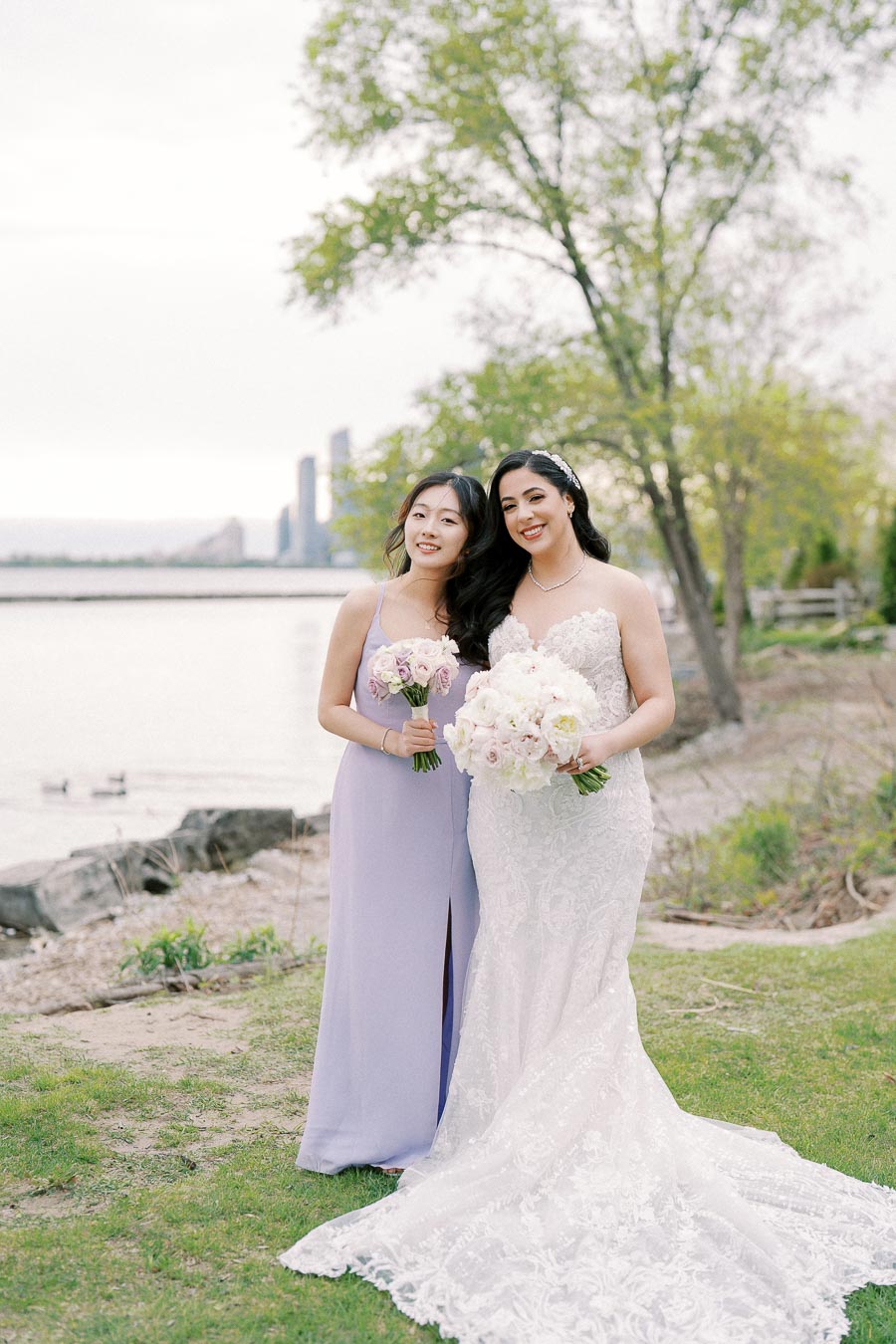 Two women pose together outdoors near a waterfront, with one in a white lace wedding dress holding a bouquet of pink and white flowers, and the other in a lilac bridesmaid dress holding a similar bouquet. The background features green trees and distant city buildings, creating a serene wedding setting.