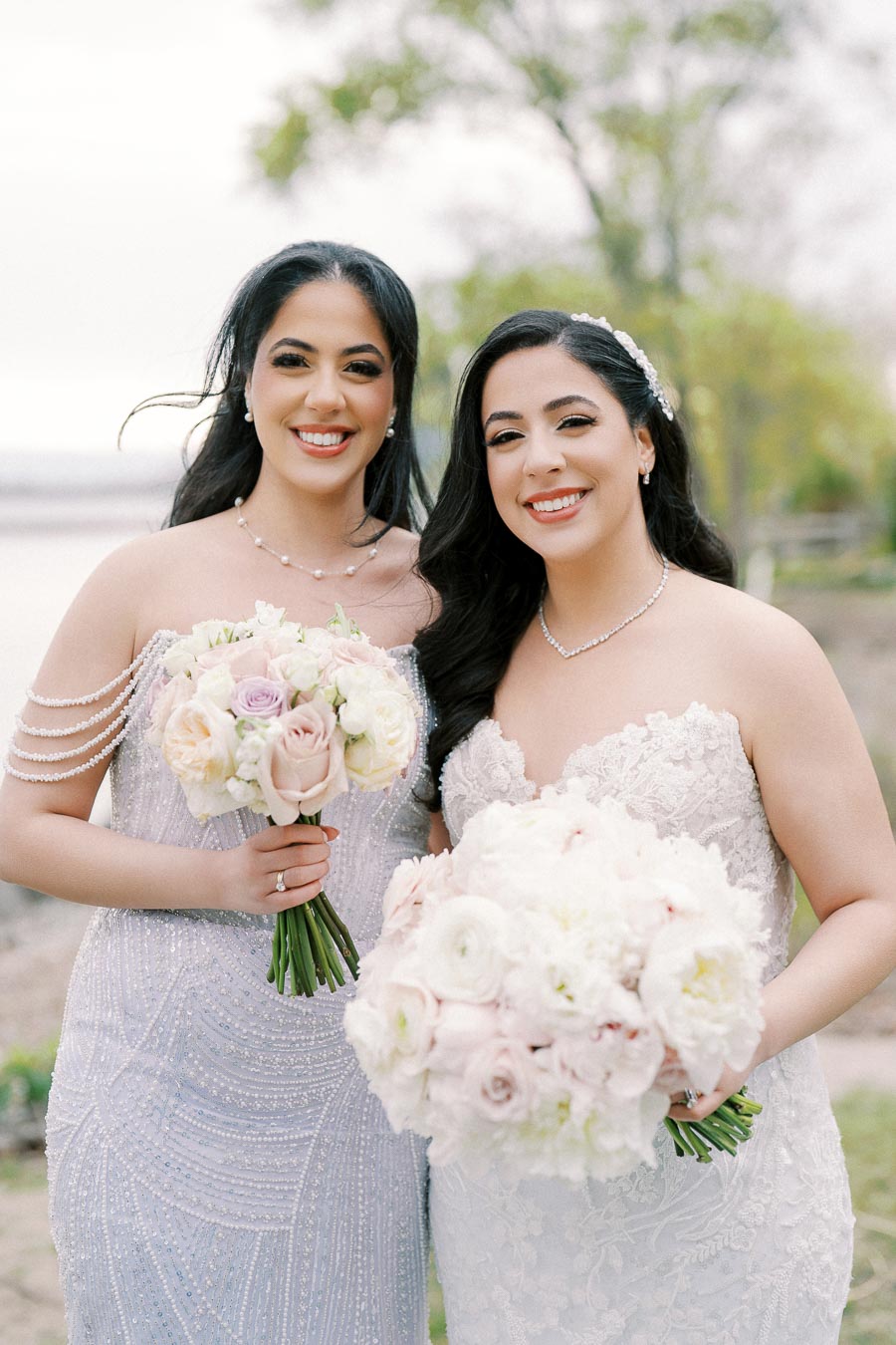 Two women in elegant lace dresses holding pastel floral bouquets, smiling outdoors with a blurred natural background, capturing a joyful moment during a wedding celebration.