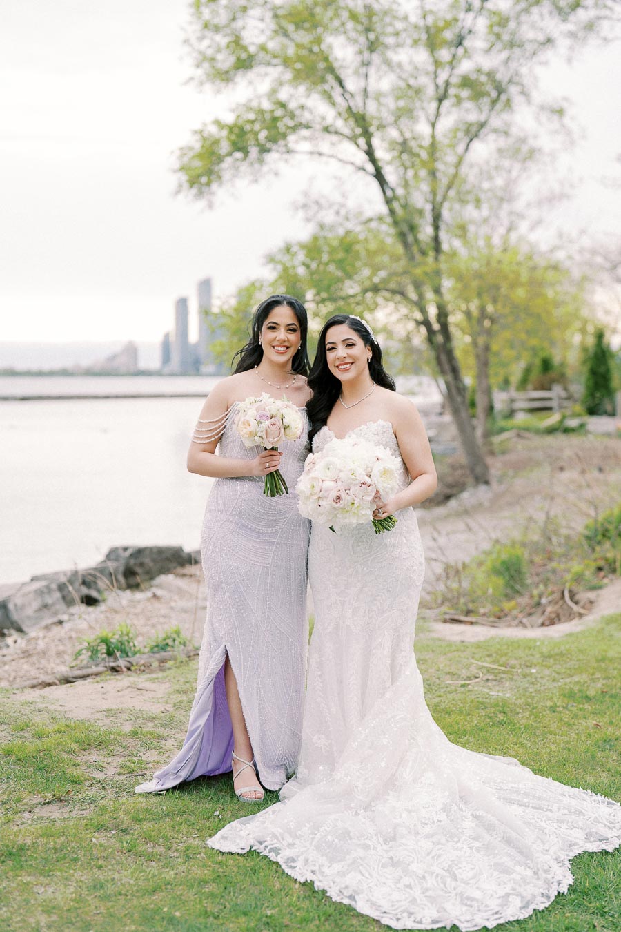 Two women in elegant dresses hold bouquets, standing outdoors by a waterside with skyscrapers in the background and surrounded by greenery.