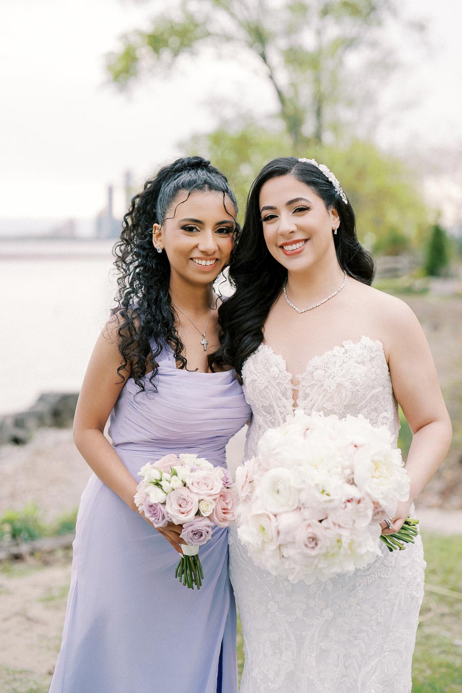 Two women smiling outdoors, one in a white lace wedding dress holding a bouquet of white and pink flowers, and the other in a lavender gown holding a similar bouquet, with a blurred natural backdrop.