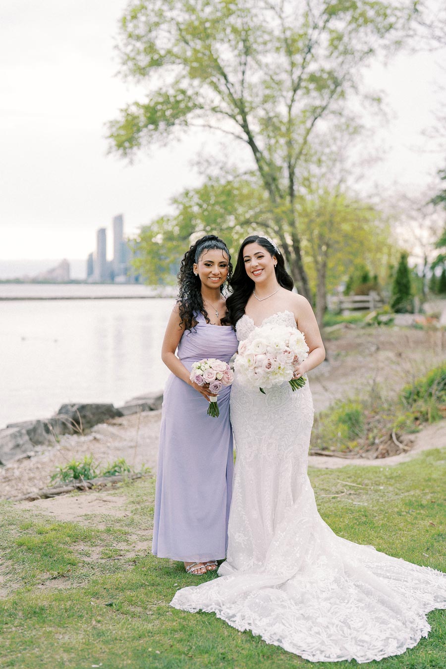 Bride in elegant white lace gown and bridesmaid in lilac dress holding bouquets, standing on a grassy waterfront with city skyline and trees in the background.