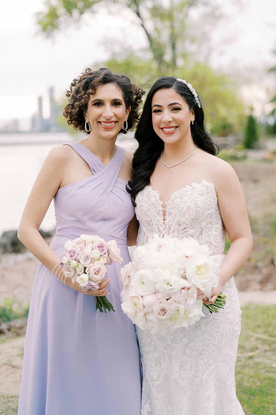 Bride and bridesmaid smiling outdoors near a waterfront, with the bride in an elegant lace gown holding a bouquet of white and pink flowers, and the bridesmaid in a lavender dress holding a matching bouquet, framed by greenery and a blurred cityscape in the background.