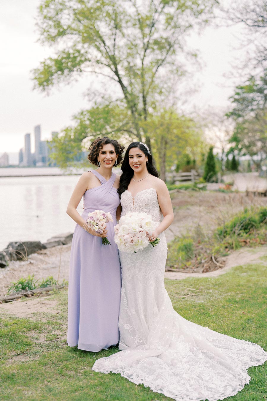 A bride in a lace wedding dress and her bridesmaid in a lavender gown stand together holding bouquets, with a scenic lakeside and city skyline in the background.