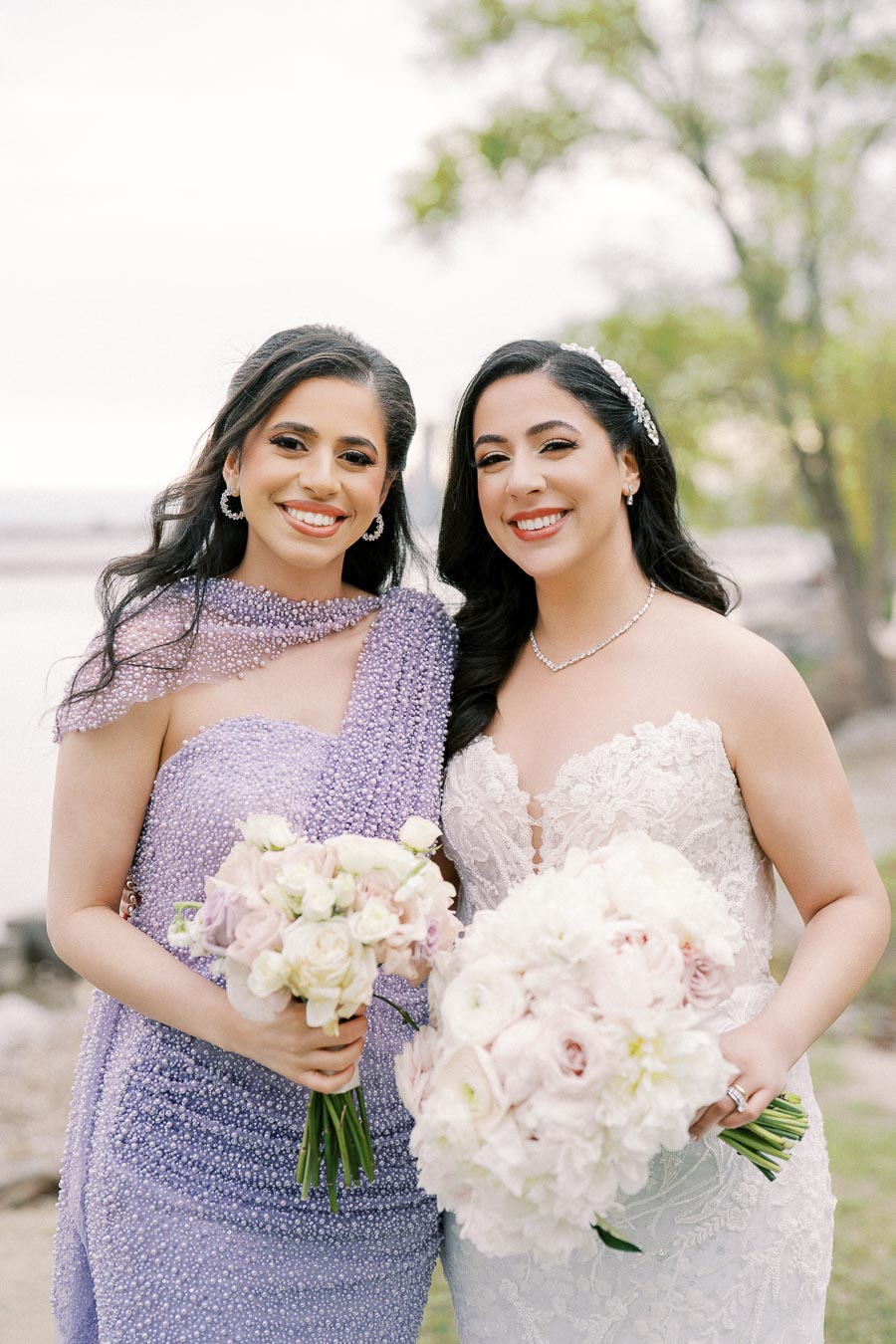 Two women smiling at an outdoor wedding, one in a lace wedding dress holding a bouquet of white and blush flowers, and the other in a sparkly lavender dress holding a matching bouquet, with a natural background featuring trees and a waterfront.