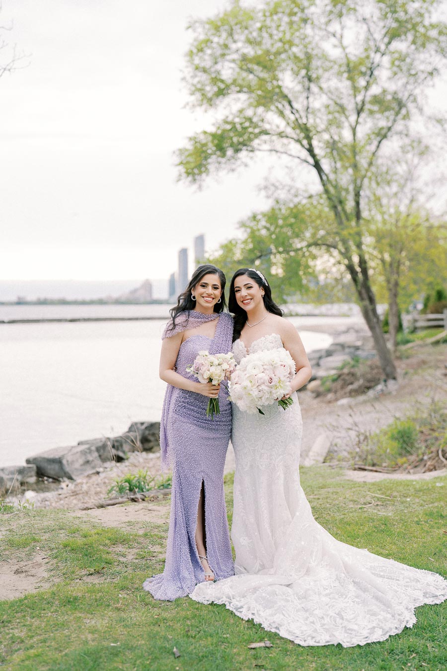 Two women in elegant dresses pose with bouquets by a waterfront, with tall buildings in the background and lush greenery surrounding them.