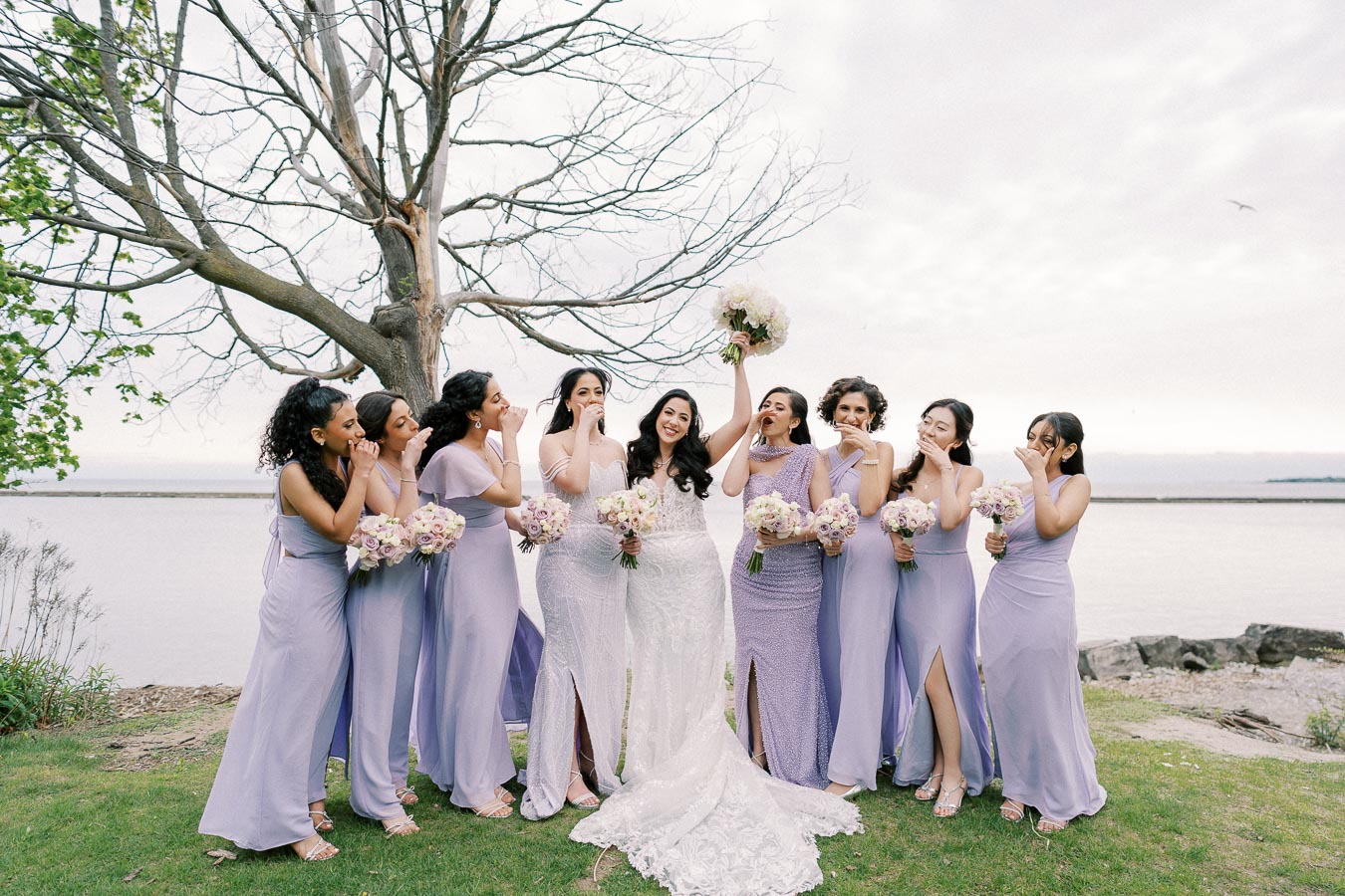 A joyful bride in a white gown stands with her bridesmaids dressed in matching lilac dresses, holding bouquets, posing near a tranquil lakeside under an overcast sky.
