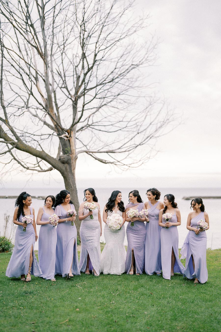 A bride with her bridesmaids in lavender dresses, holding bouquets, standing outdoors near a leafless tree by a waterfront.