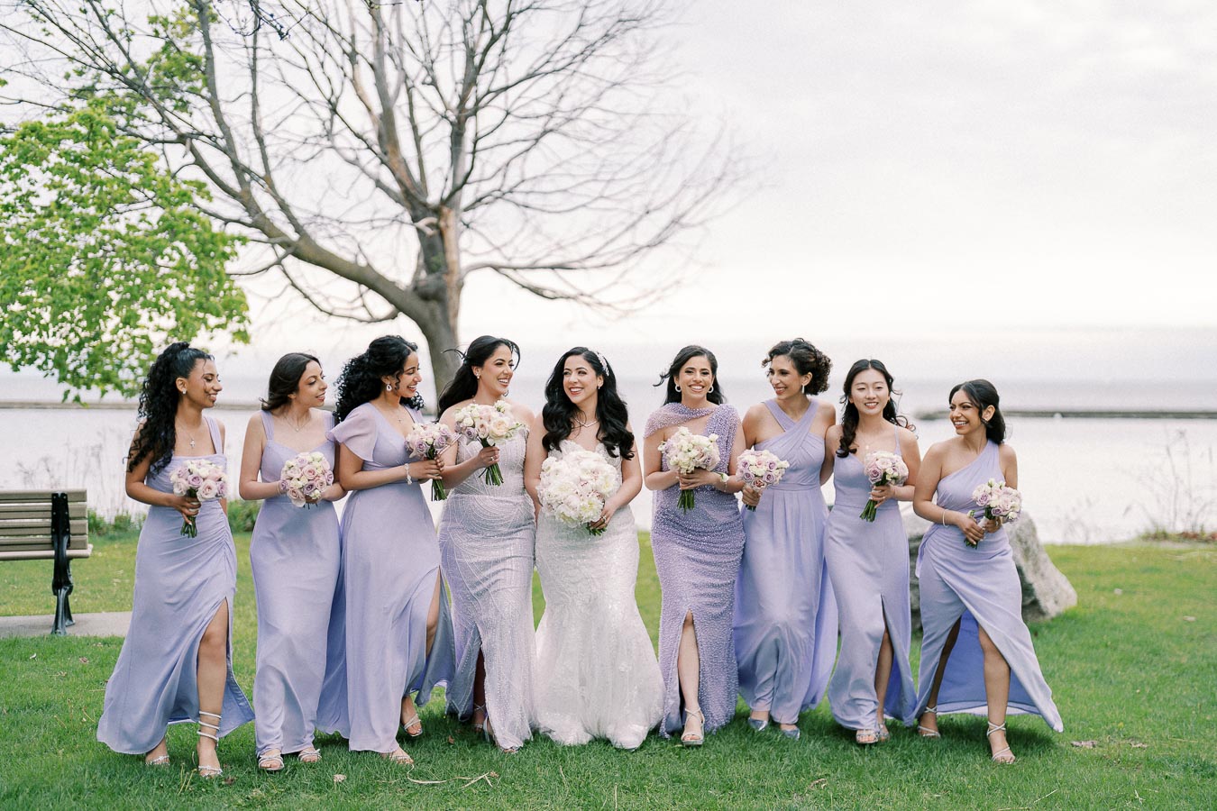 Bridal party in lavender dresses with bouquets walking on grass near a tree and waterfront.