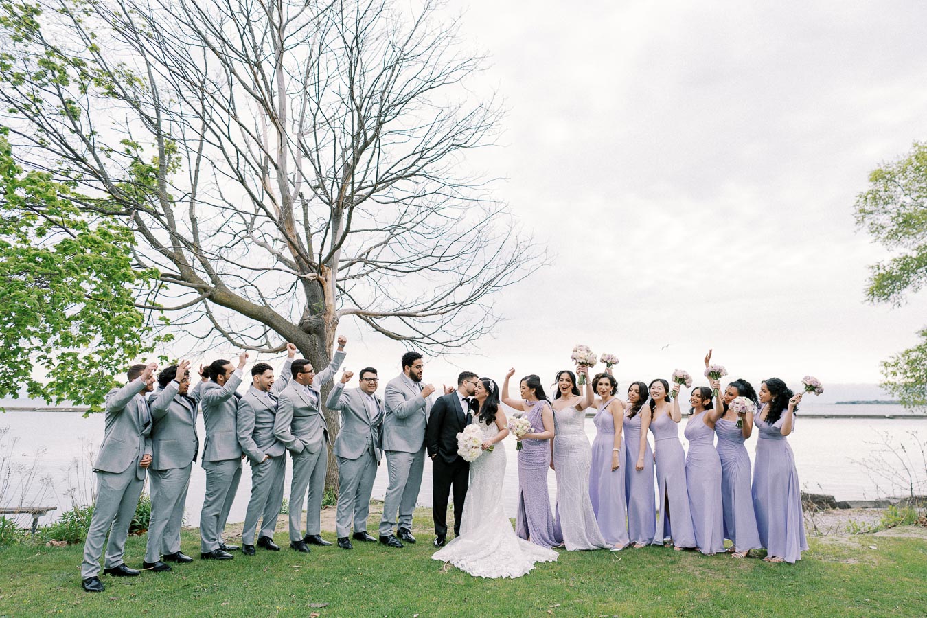 A joyful wedding party poses on a grassy lakeside, featuring a bride in a white gown and a groom in a black suit, surrounded by bridesmaids in lavender dresses and groomsmen in gray suits, under a large tree with budding leaves and a scenic water backdrop.