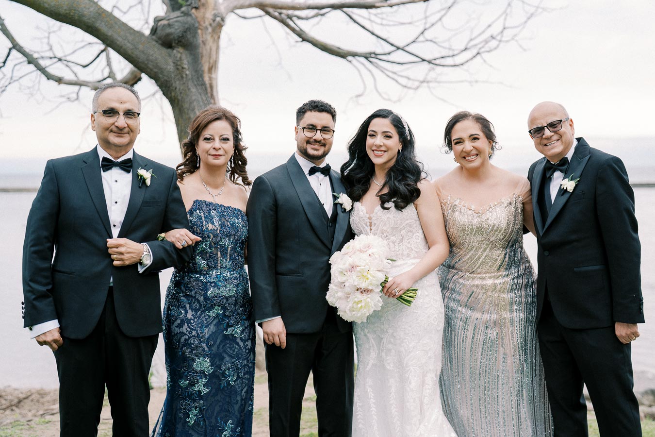 Wedding group photo featuring elegantly dressed couple and family members in formal attire posing by a tree near a waterfront. The bride holds a bouquet of white flowers, and everyone is smiling.