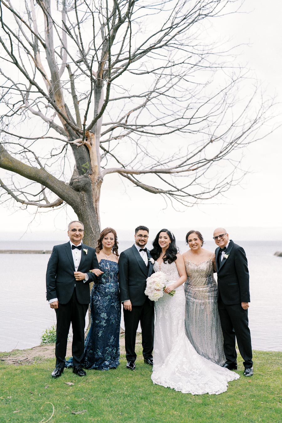 A wedding group photo with six people elegantly dressed, standing by a bare tree near a waterfront. The bride holds a bouquet of roses, wearing a white lace gown, while the guests wear formal suits and evening dresses, celebrating the occasion outdoors on a grassy area.