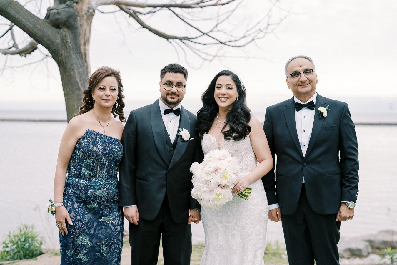 A bride and groom pose with two guests in formal attire by a scenic waterfront, featuring greenery and a bare tree in the background. The bride holds a bouquet of white and pink flowers.